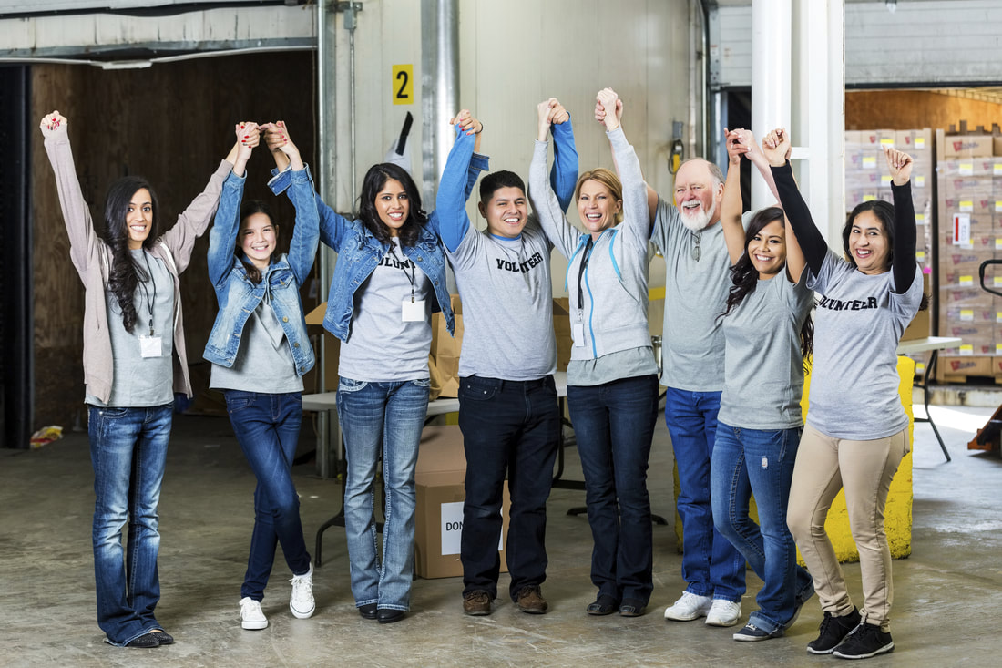 A group of eight volunteers at a food pantry sorting facility who have all joined hands and raised them above their heads in a happy cheer. All of them are wearing tee shirts that say 
